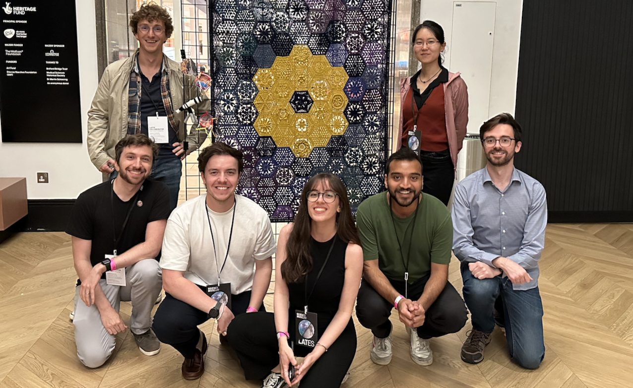 A group of Astronomy PhDs and postdocs at the Science Museum, posing in front of a crochet of the JWST mirror
