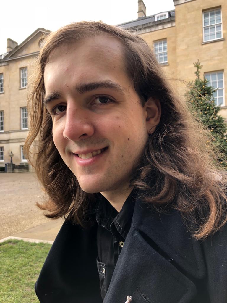 A smiling person with long hair, dressed in a black trenchcoat and shirt, in front of historic buildings.